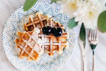 Waffles on a blue plate, topped with red cherries. Fork and knife off to the side