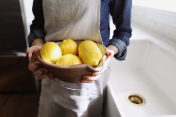Photo of a woman in an apron holding a bowl full of lemons while standing next to a sink in a kitchen