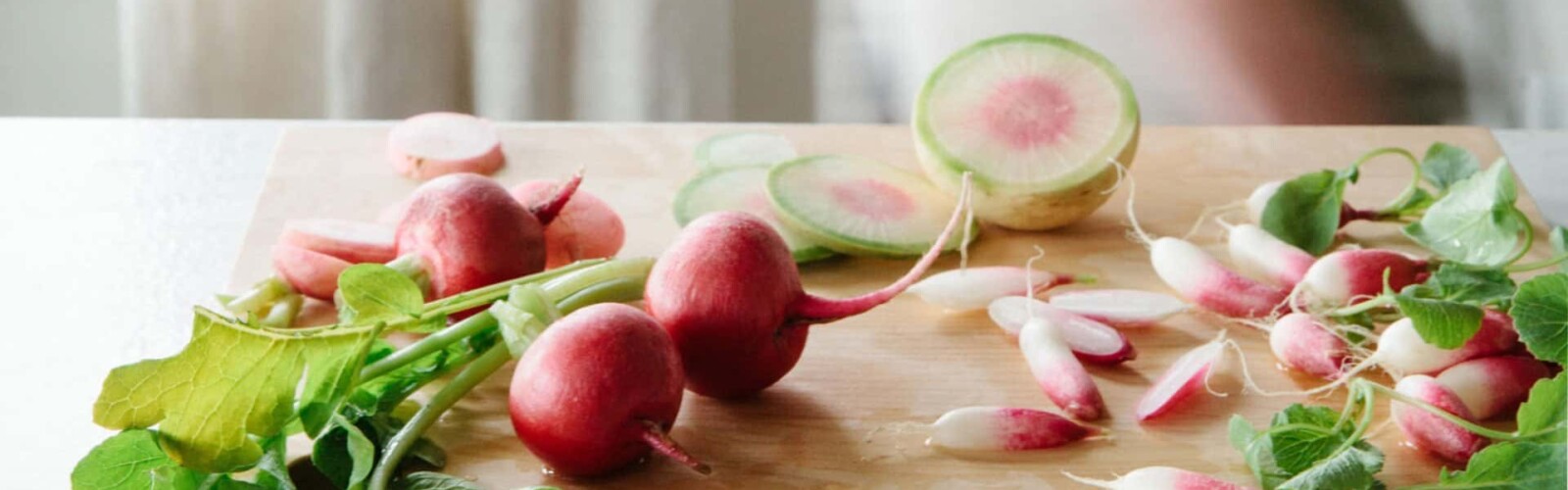 Red radishes on a wood cutting board, with a woman walking by through the kitchen