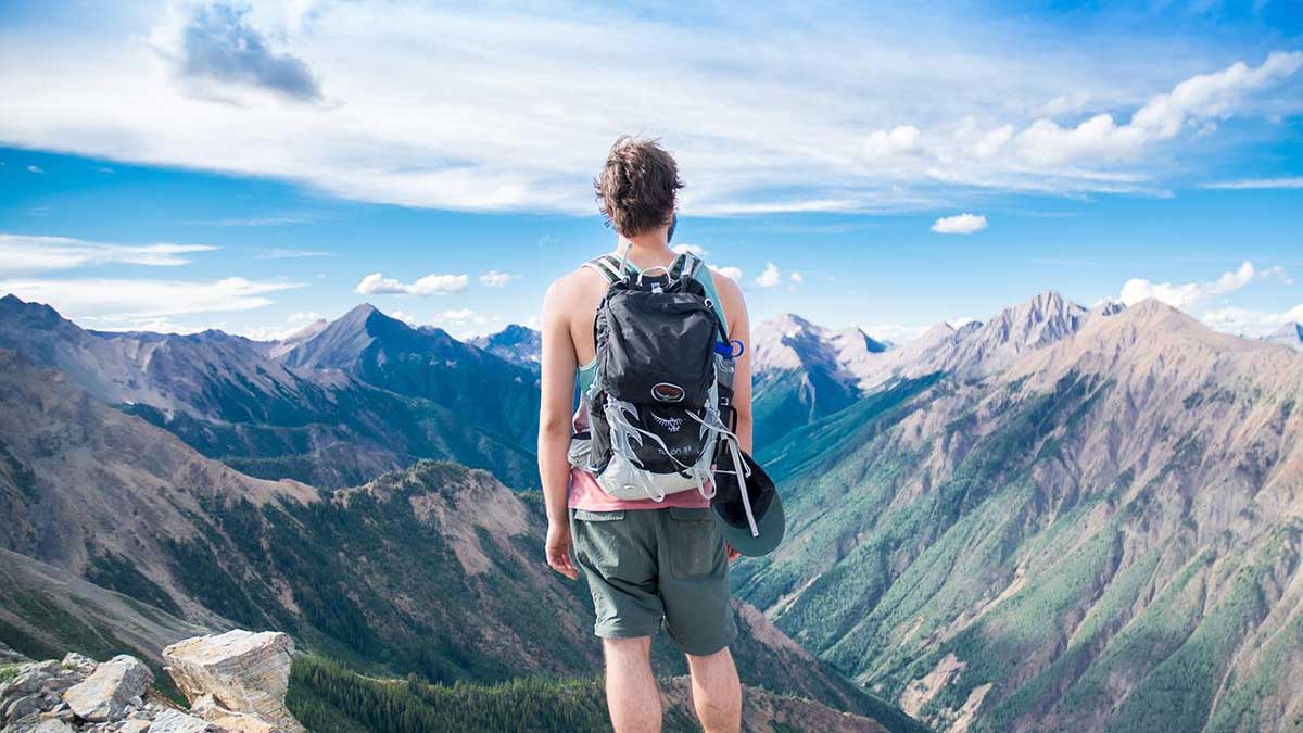 hiker stands on mountain top looking at bright blue skies and deep blue mountains.